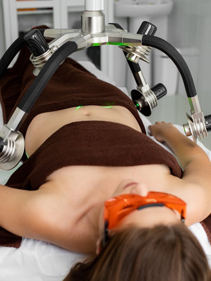 Services Female patient covered with towels in protective eyeshades lying under modern laser weight loss machine during treatment in medical clinic